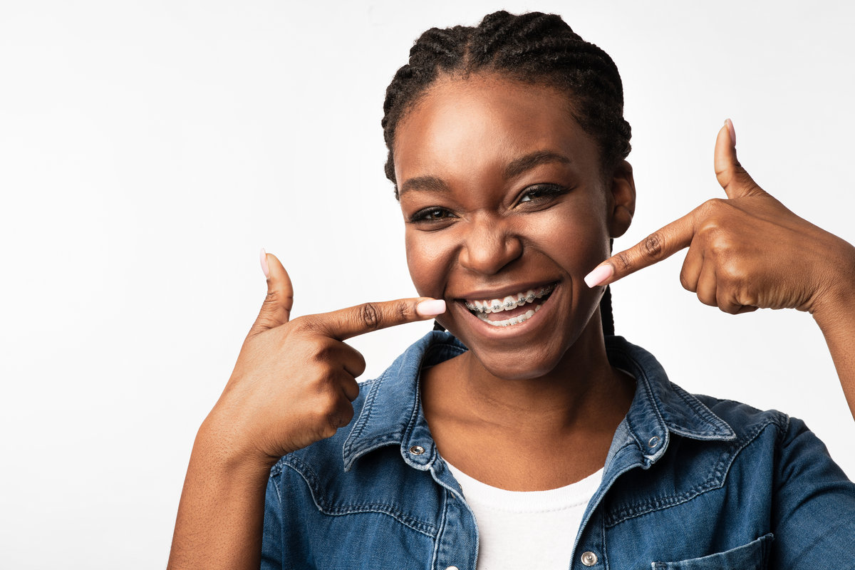 Joyful Black Girl In Orthodontic Braces Smiling Pointing Fingers At Brackets On Her Teeth Posing Over White Background. Studio Shot