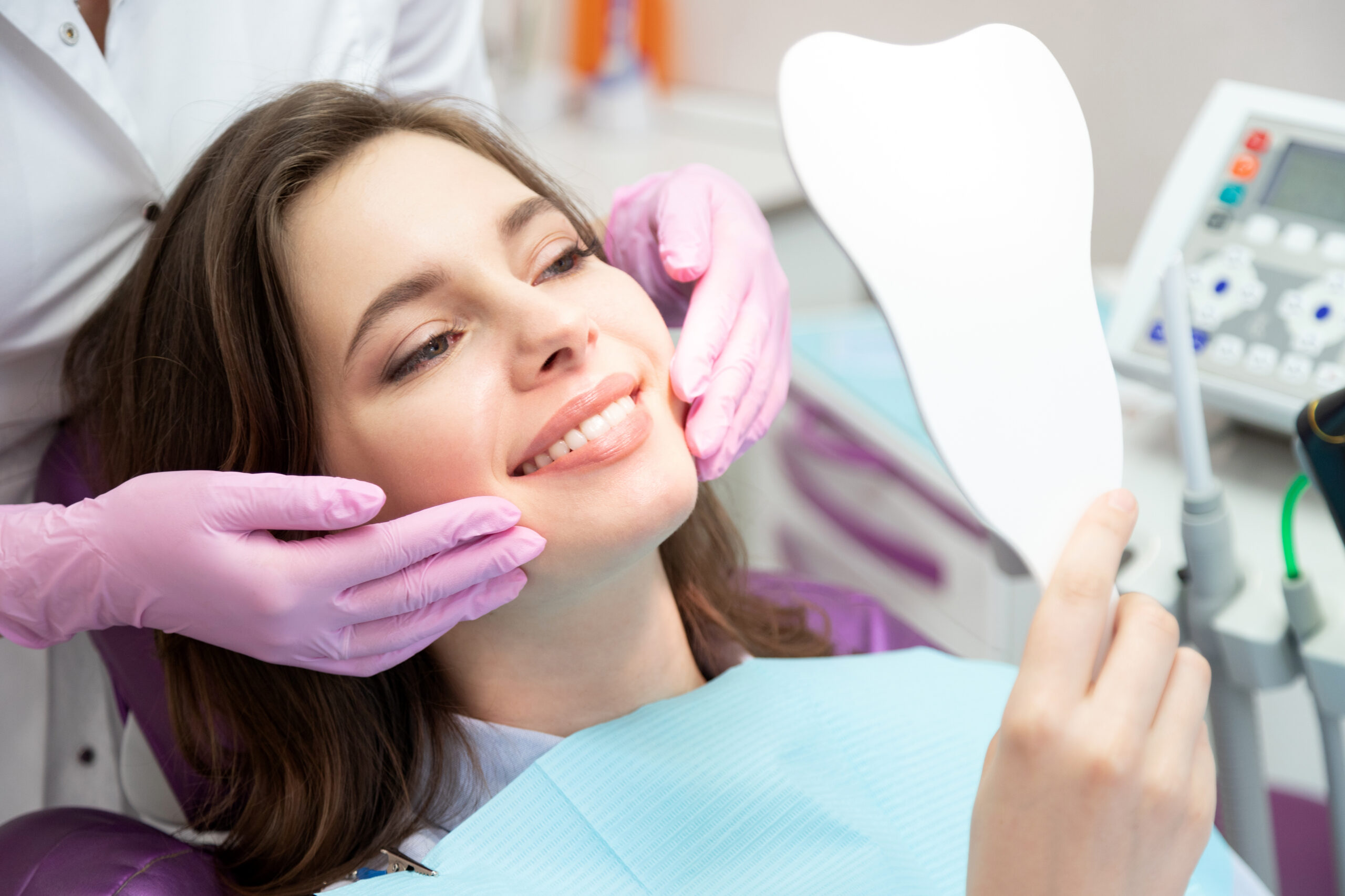 Young woman in dental chair smiling while dentist in pink gloves examines her teeth