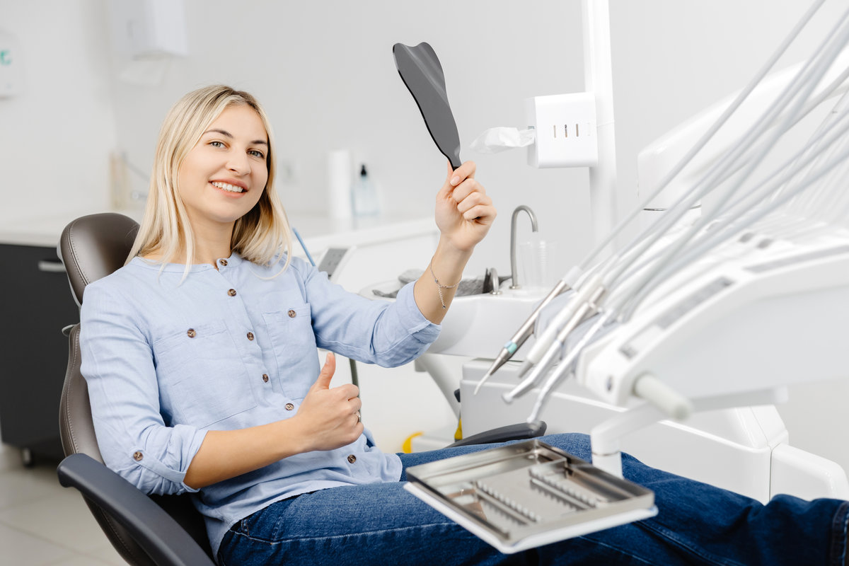 Smiling patient is checking her teeth after dental procedure using small mirror in modern dental clinic
