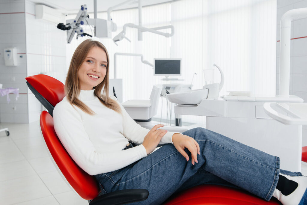 A beautiful young woman is sitting on a red dental chair in modern white dentistry and smiling. Dental prosthetics, treatment and teeth whitening