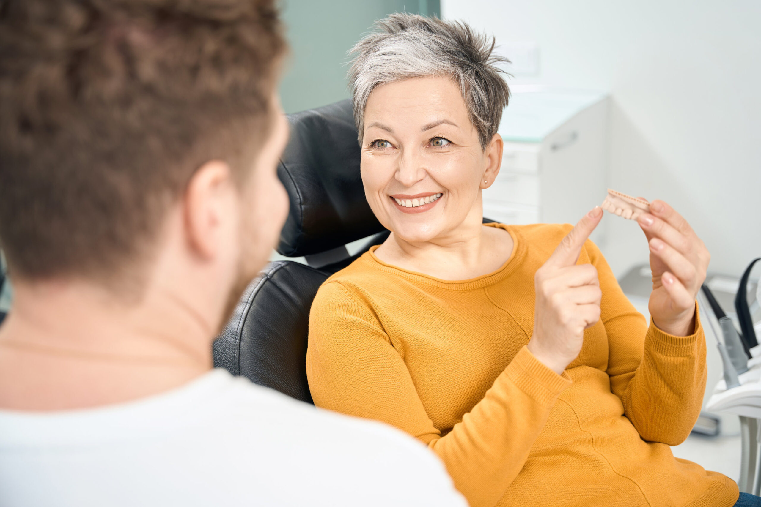Positive adult woman client of aesthetic dentistry clinic visiting doctor office to discuss the theme of dental implants or dentures, holding in hands 3d teeth model